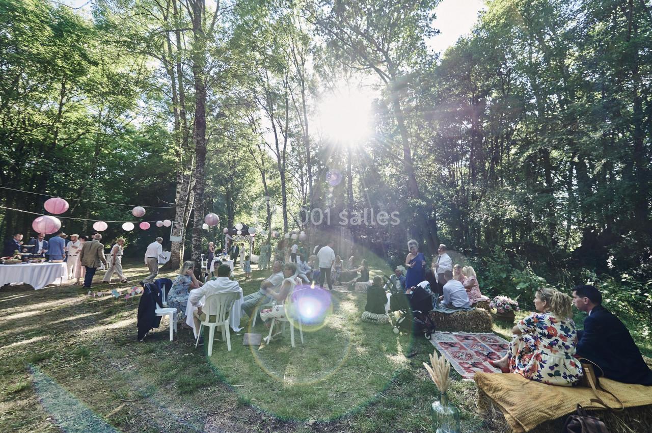 Des invités assis et debout participent à une réception en plein air dans une clairière ensoleillée décorée de lanternes.