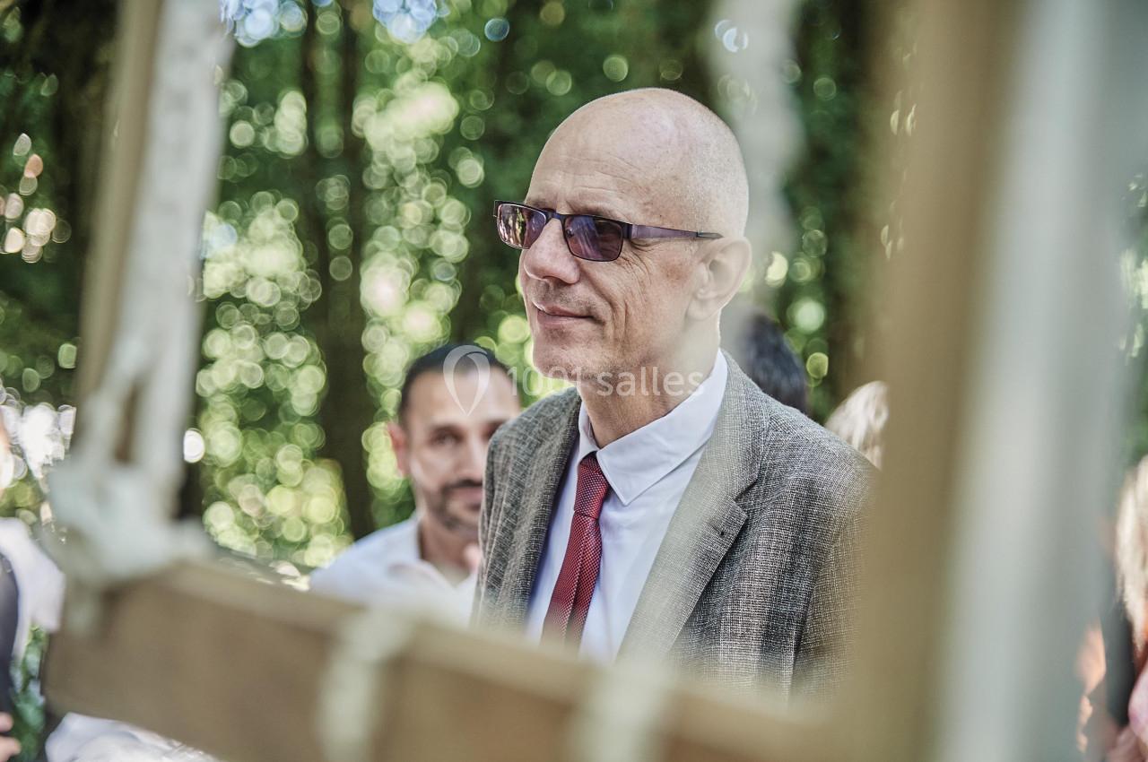 Un homme chauve en costume et cravate rouge, portant des lunettes, se tient dans un environnement boisé.