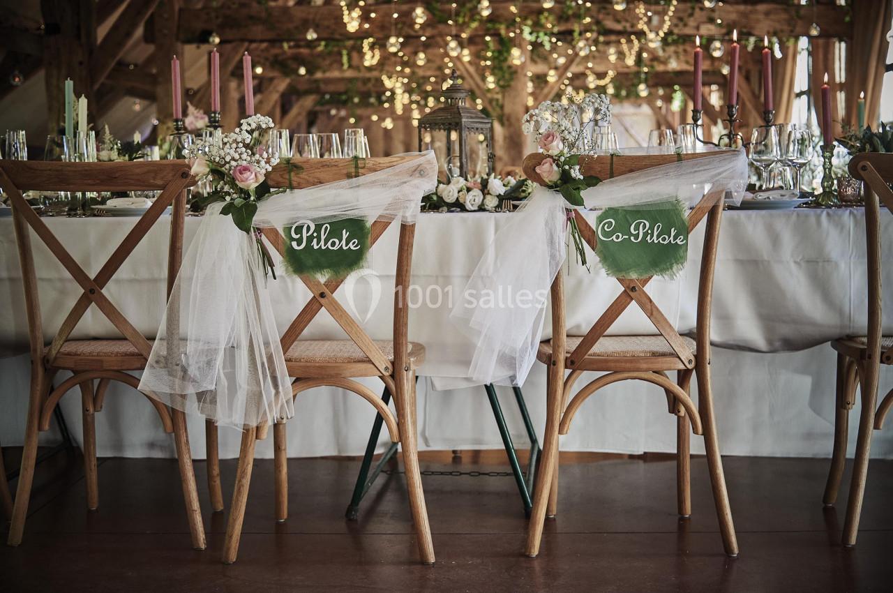 Chaises en bois décorées de voilages et de panneaux ’Pilote’ et ’Co-Pilote’ devant une table de réception ornée de fleurs.