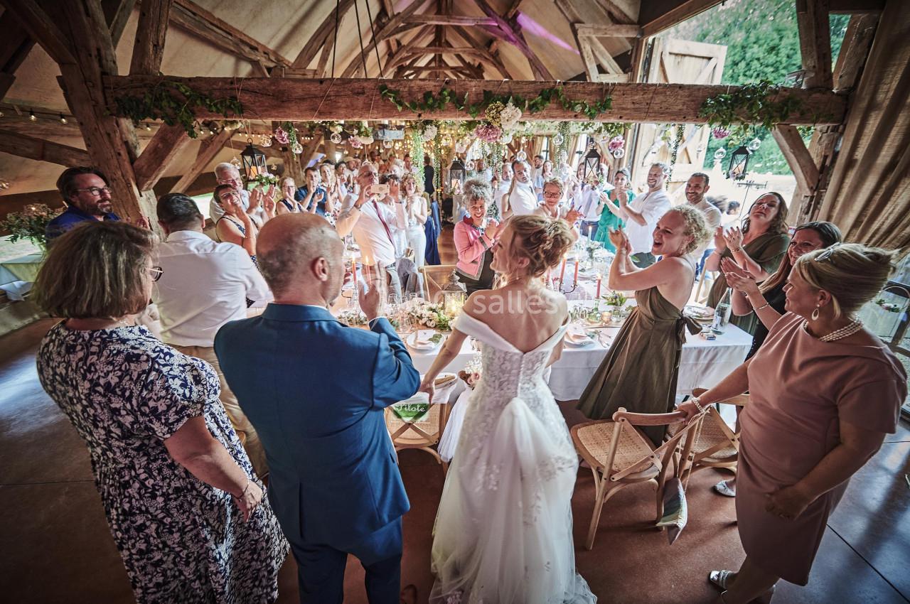 Un couple de mariés debout dans une salle en bois, entouré d'invités applaudissant lors d'une réception.