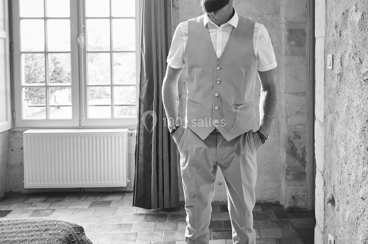 Un homme debout dans une pièce lumineuse avec un sol en pierre, portant un gilet et une chemise.