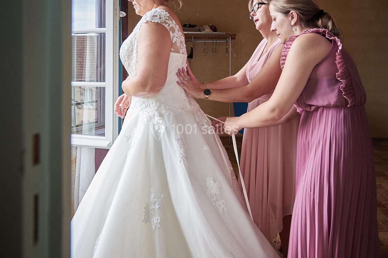 Deux femmes en robes roses ajustent la robe de mariée d'une femme près d'une fenêtre dans une pièce lumineuse.