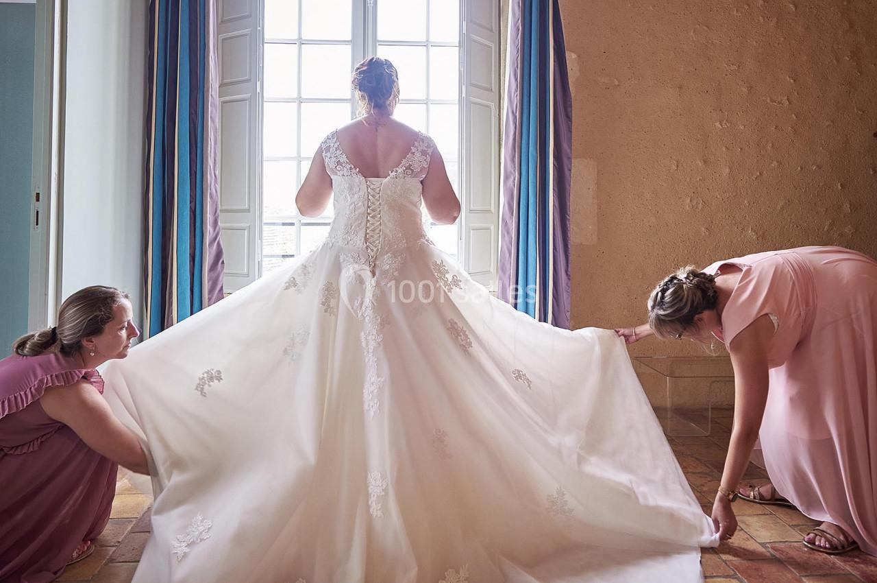 Une mariée en robe blanche est aidée par deux femmes à ajuster sa tenue près d'une fenêtre lumineuse.