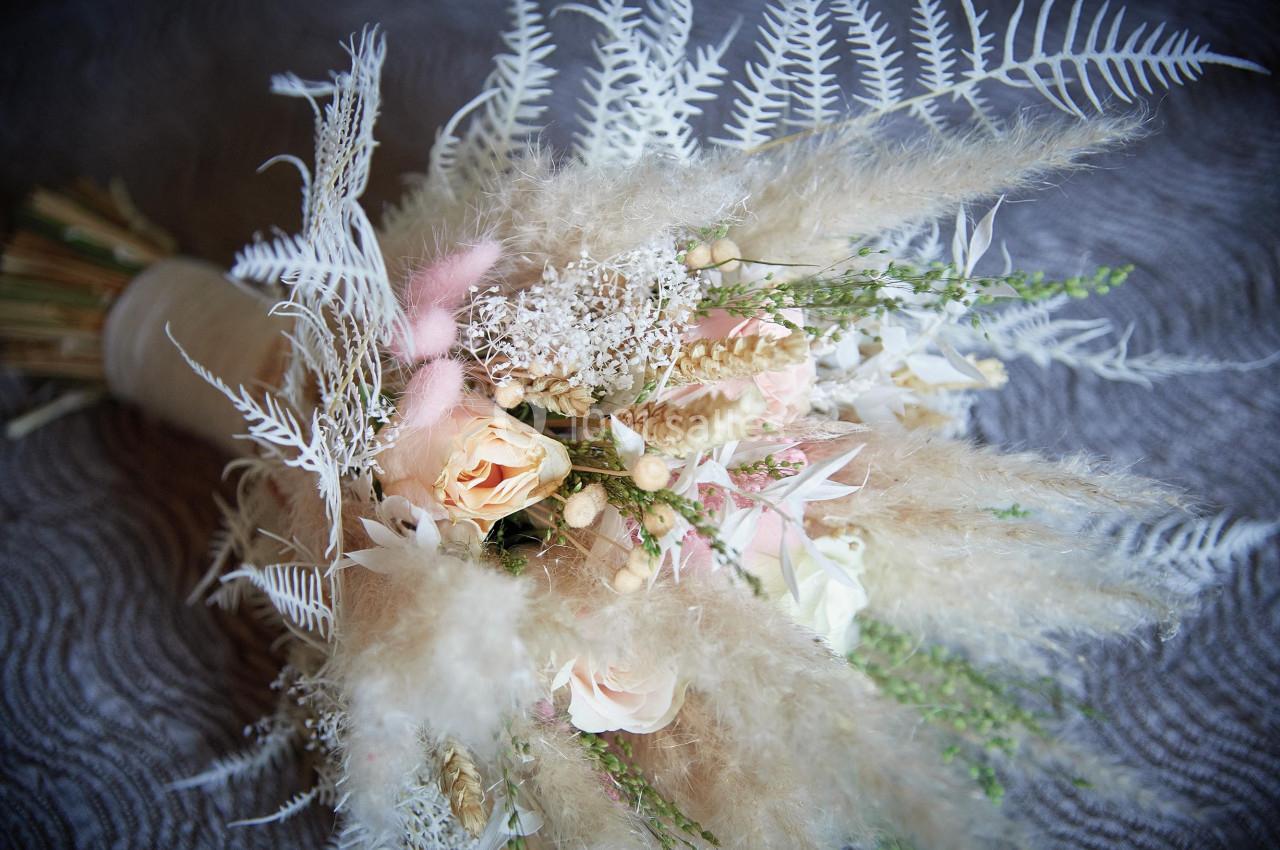 Bouquet de fleurs séchées avec des roses pâles, des herbes et des fougères blanches sur un fond texturé gris.