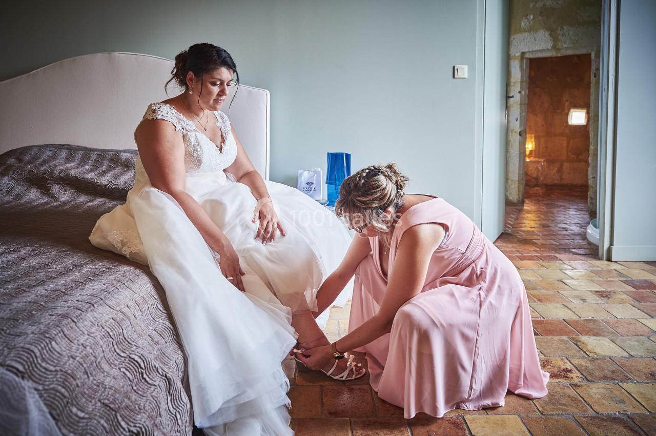Une femme en robe de mariée assise sur un lit, aidée par une autre femme à ajuster ses chaussures.