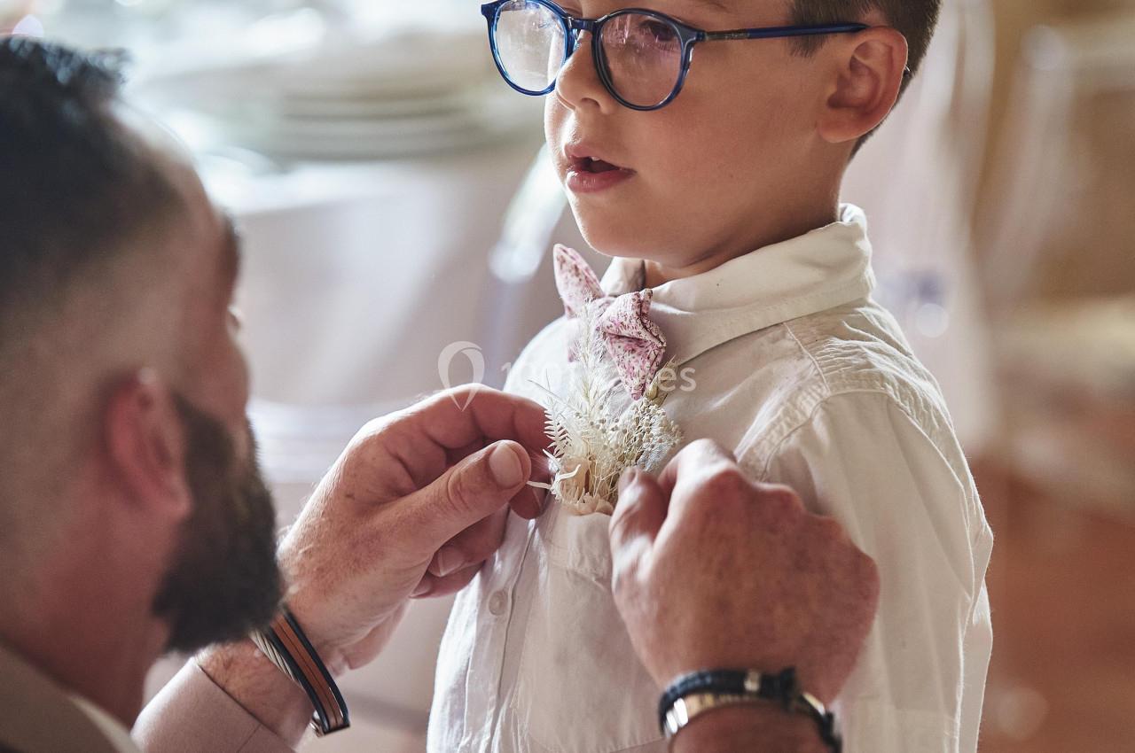 Un homme ajuste une boutonnière sur la chemise d'un jeune garçon portant des lunettes et un nœud papillon.