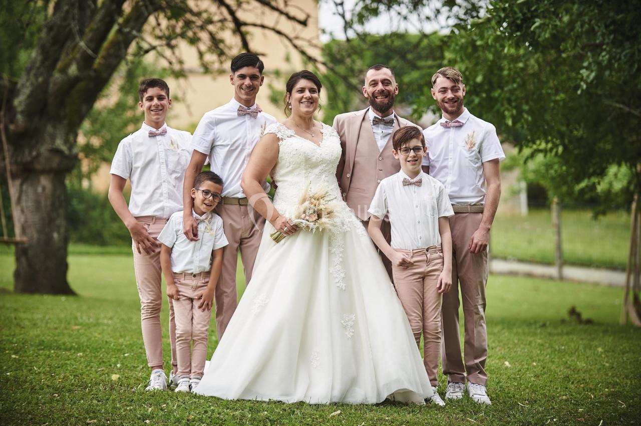 Un couple en tenue de mariage pose avec cinq garçons habillés en chemises blanches et pantalons beige dans un jardin.
