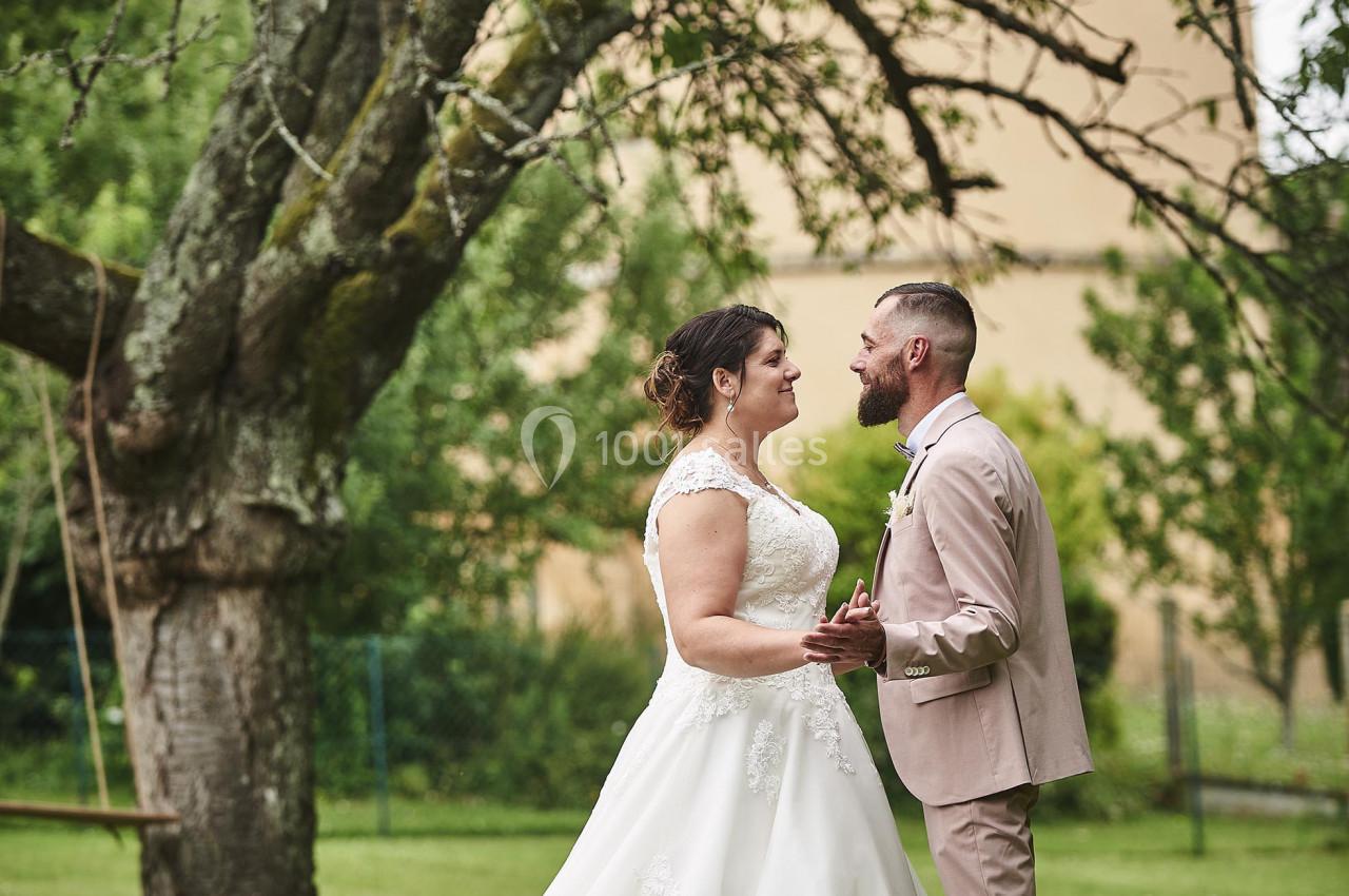 Un couple en tenue de mariage se tient face à face dans un jardin verdoyant près d'un grand arbre.