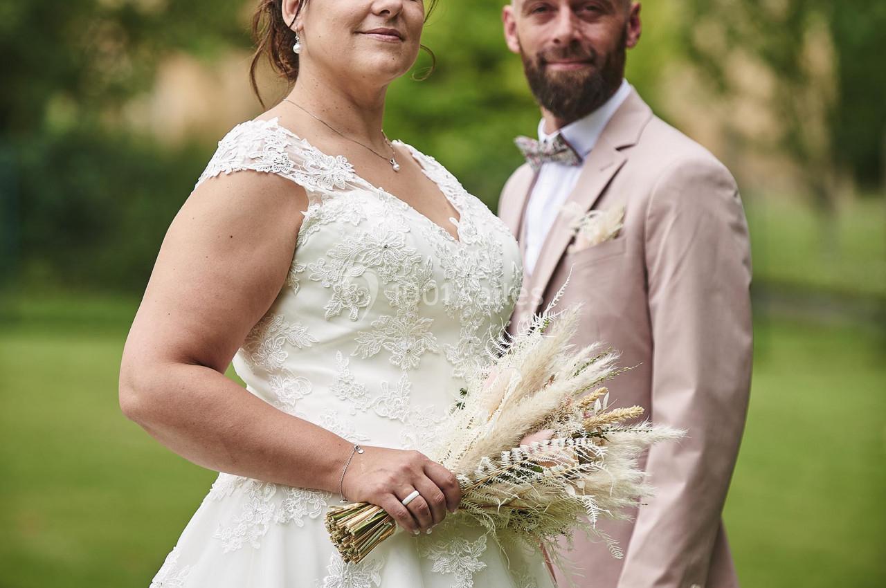 Un couple en tenue de mariage pose dans un jardin, la mariée tenant un bouquet de fleurs séchées.