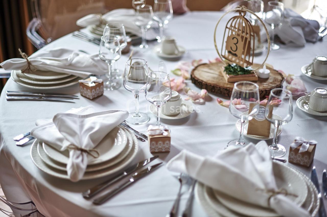 Table élégamment dressée avec nappes blanches, vaisselle raffinée, verres, serviettes pliées et décoration centrale en bois.