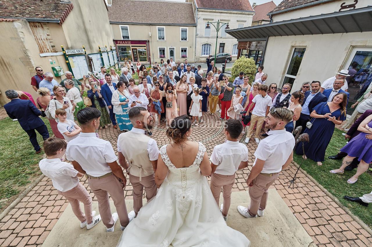 Un couple de mariés et leurs témoins font face à une assemblée réunie en extérieur devant des bâtiments.