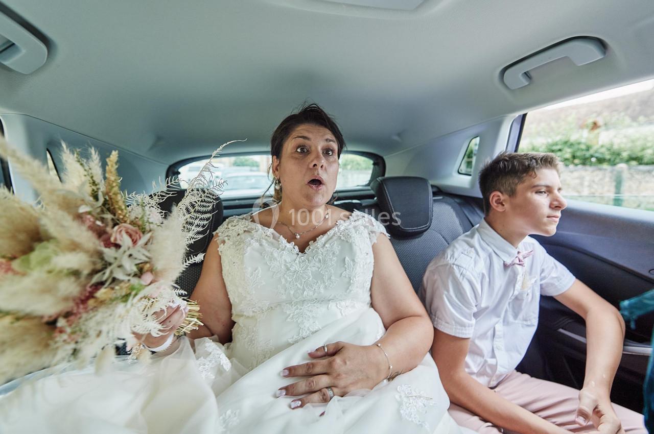 Une femme en robe de mariée et un jeune garçon assis à l'arrière d'une voiture, tenant un bouquet de fleurs séchées.