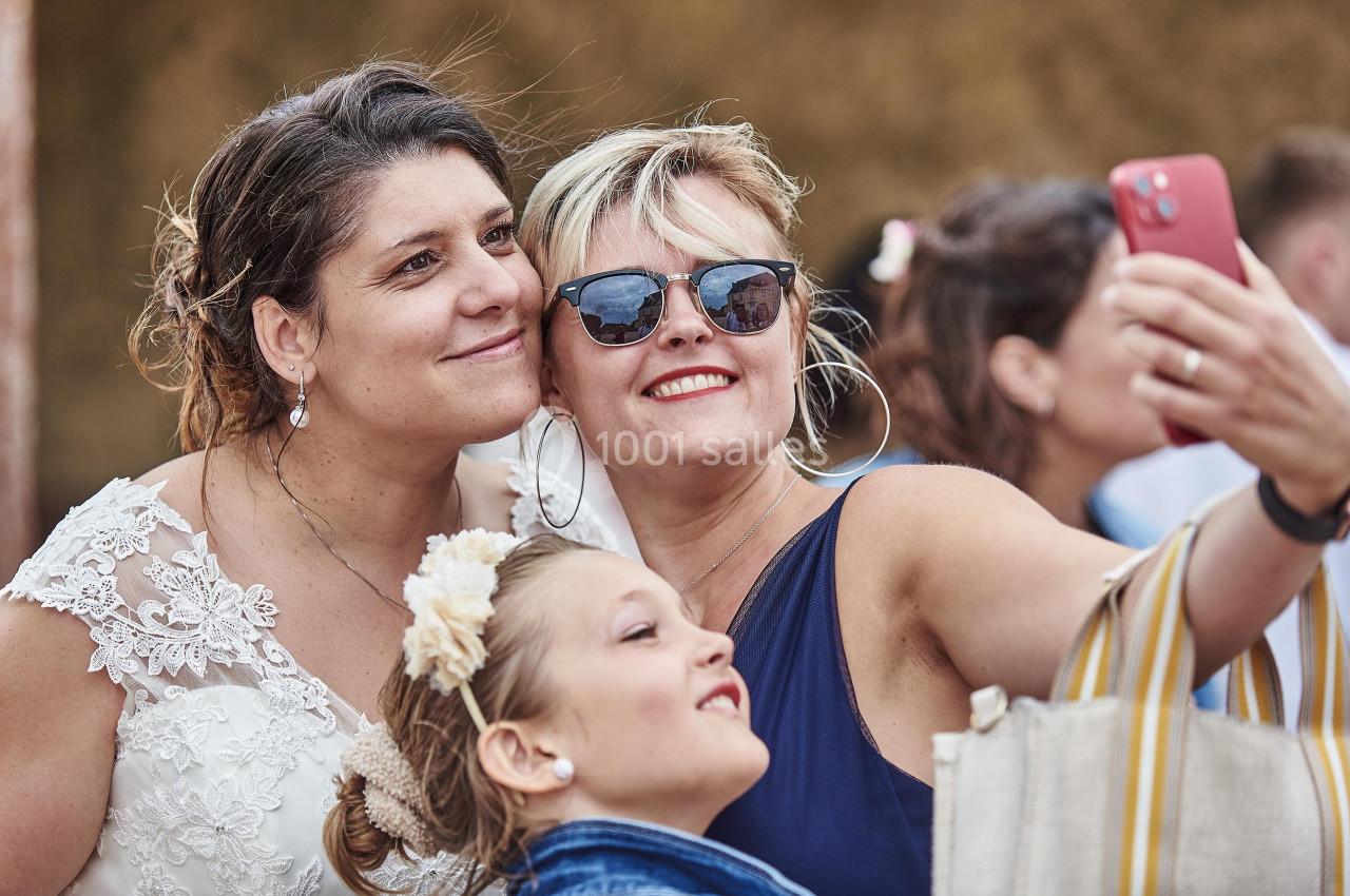 Deux femmes souriantes, dont une en robe de mariée, prennent un selfie avec une jeune fille.