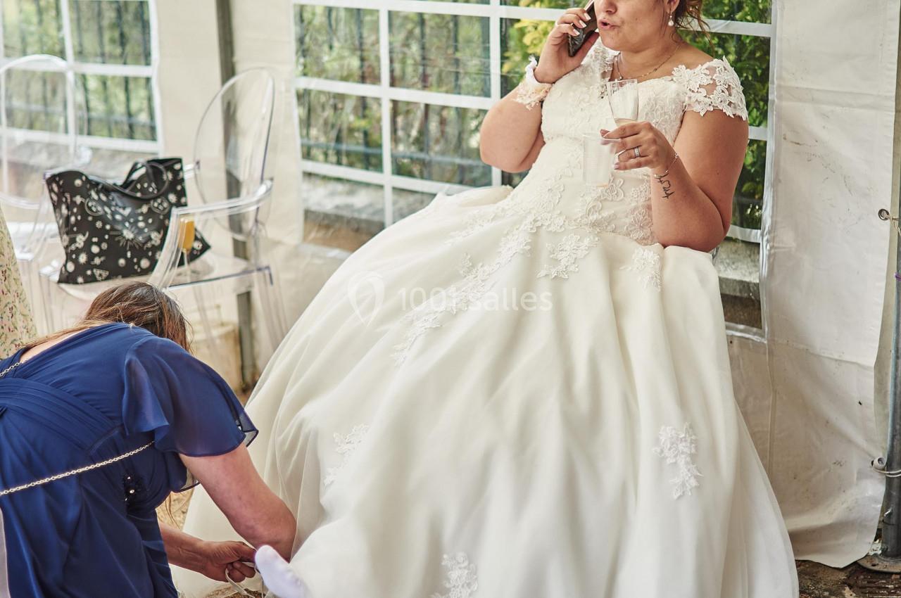 Une mariée en robe blanche parle au téléphone tandis qu'une femme ajuste sa chaussure sous une tente.