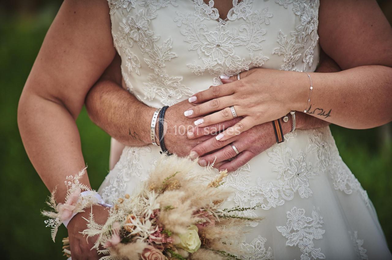 Un couple en tenue de mariage se tient par la taille, avec un bouquet de fleurs séchées au premier plan.