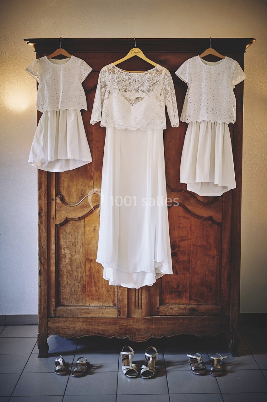 Trois robes blanches, dont une de mariée, suspendues à une armoire en bois avec des chaussures posées au sol.