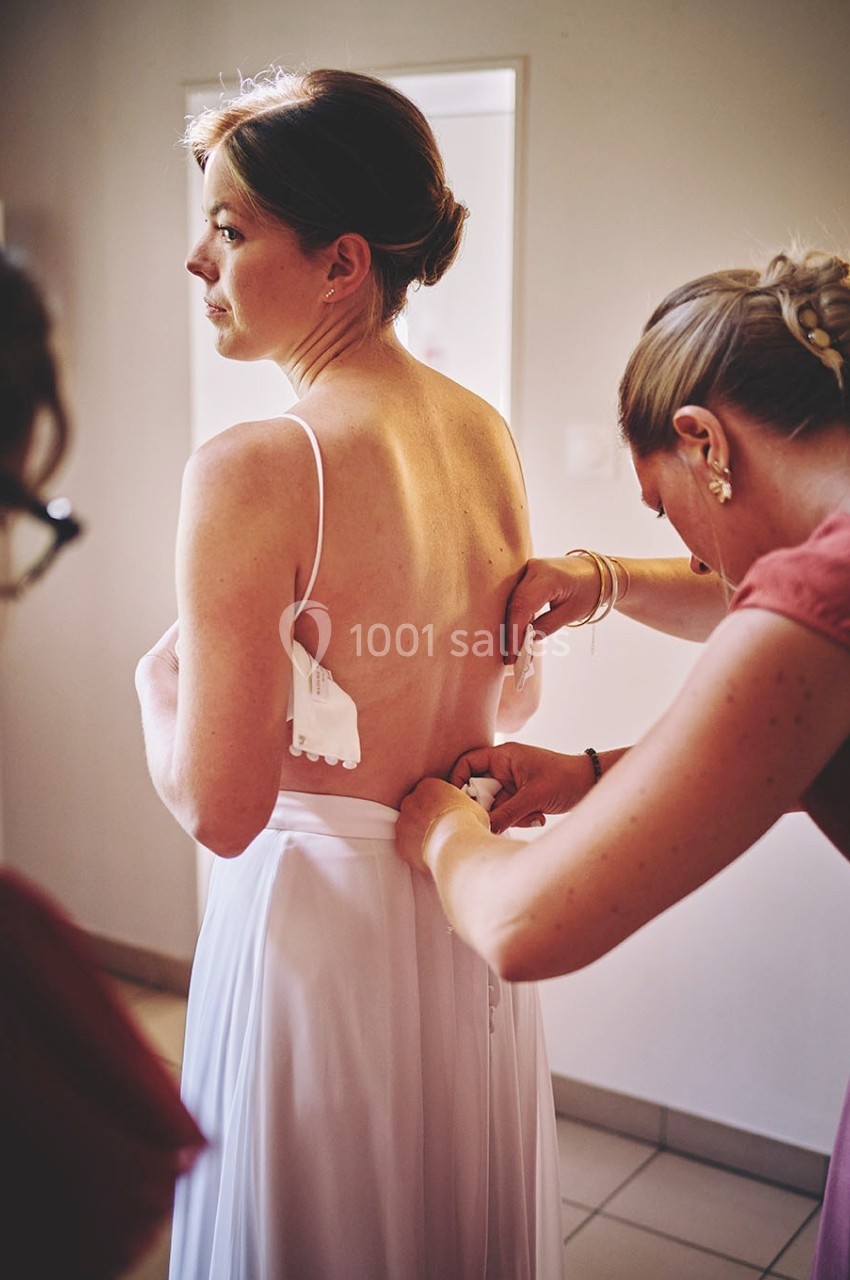 Une femme en robe blanche est aidée par une autre personne pour ajuster le dos de sa tenue dans une pièce éclairée.