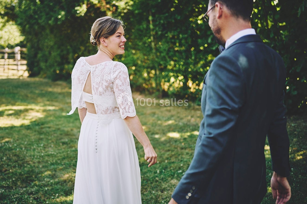 Une femme en robe de mariée marche dans un jardin, suivie par un homme en costume.