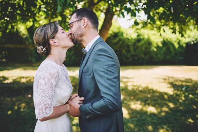 Un couple de mariés s'embrasse tendrement près d'un étang, entouré de verdure, la mariée tenant un bouquet coloré.