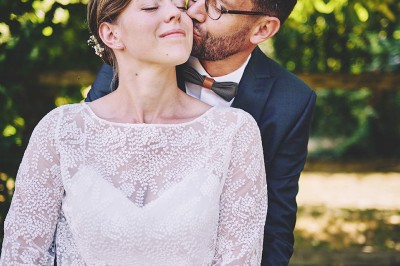 Un couple de mariés s'embrasse tendrement près d'un étang, entouré de verdure, la mariée tenant un bouquet coloré.