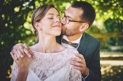 Un couple de mariés s'embrasse tendrement près d'un étang, entouré de verdure, la mariée tenant un bouquet coloré.