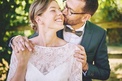 Un couple de mariés s'embrasse tendrement près d'un étang, entouré de verdure, la mariée tenant un bouquet coloré.