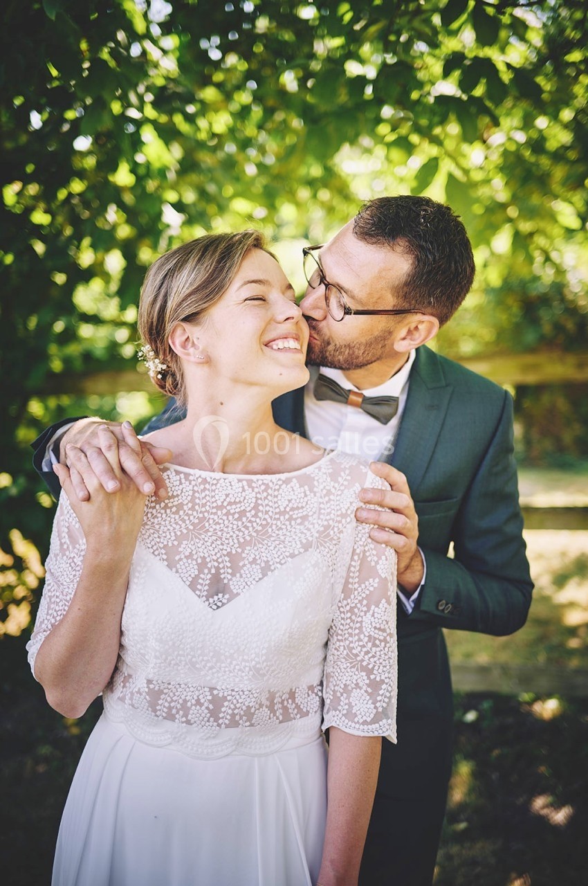 Un couple en tenue de mariage, l'homme embrassant la femme sur la joue dans un jardin ensoleillé.