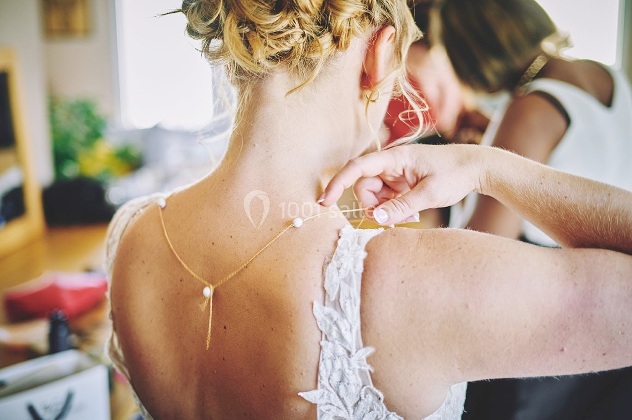 Une femme ajuste un collier délicat avec des perles sur la nuque d'une mariée portant une robe en dentelle.