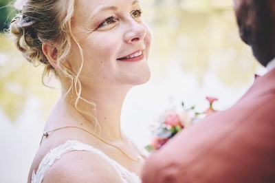 Un couple de mariés s'embrasse tendrement près d'un étang, entouré de verdure, la mariée tenant un bouquet coloré.