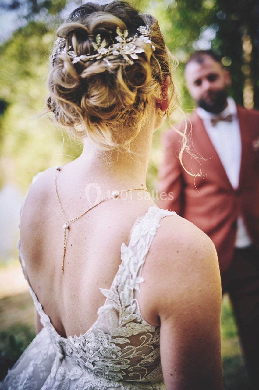 Une femme en robe de mariée vue de dos, coiffée d'un chignon orné, face à un homme en costume dans un cadre naturel.