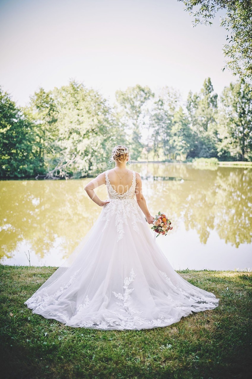 Une mariée en robe blanche élégante se tient de dos devant un lac entouré de verdure.