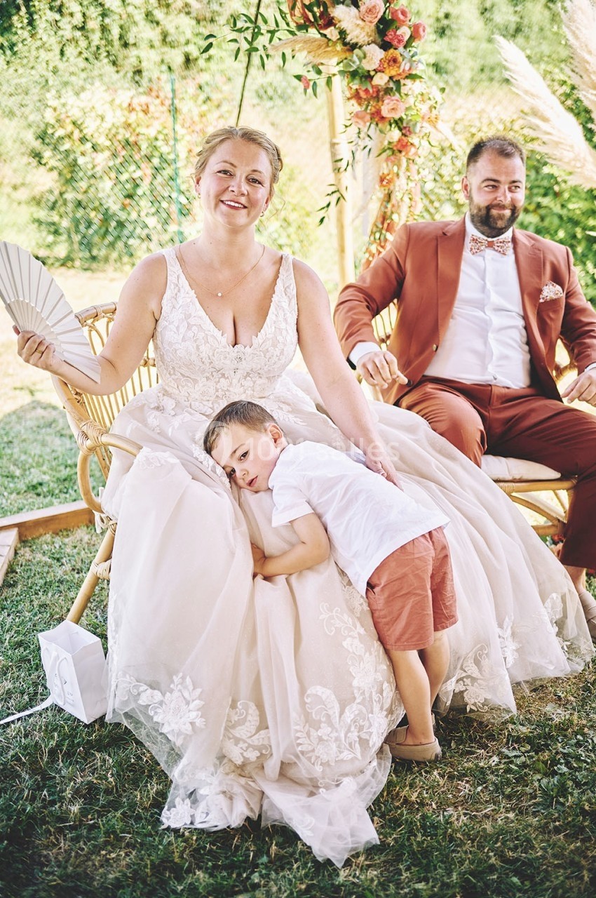 Une femme en robe de mariée assise avec un éventail, un enfant appuyé sur elle, et un homme en costume à ses côtés.