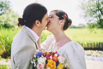 Un couple de mariés s'embrasse tendrement près d'un étang, entouré de verdure, la mariée tenant un bouquet coloré.