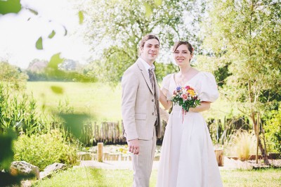 Un couple de mariés s'embrasse tendrement près d'un étang, entouré de verdure, la mariée tenant un bouquet coloré.