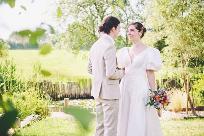 Un couple de mariés s'embrasse tendrement près d'un étang, entouré de verdure, la mariée tenant un bouquet coloré.