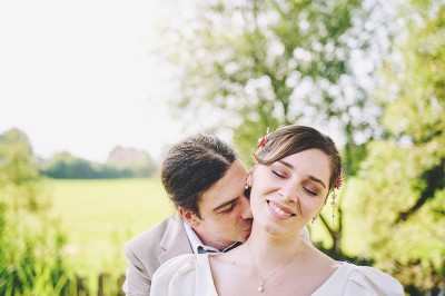 Un couple de mariés s'embrasse tendrement près d'un étang, entouré de verdure, la mariée tenant un bouquet coloré.