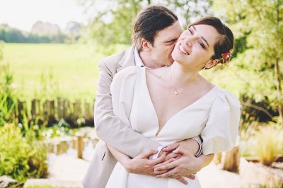 Un couple de mariés s'embrasse tendrement près d'un étang, entouré de verdure, la mariée tenant un bouquet coloré.