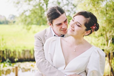 Un couple de mariés s'embrasse tendrement près d'un étang, entouré de verdure, la mariée tenant un bouquet coloré.