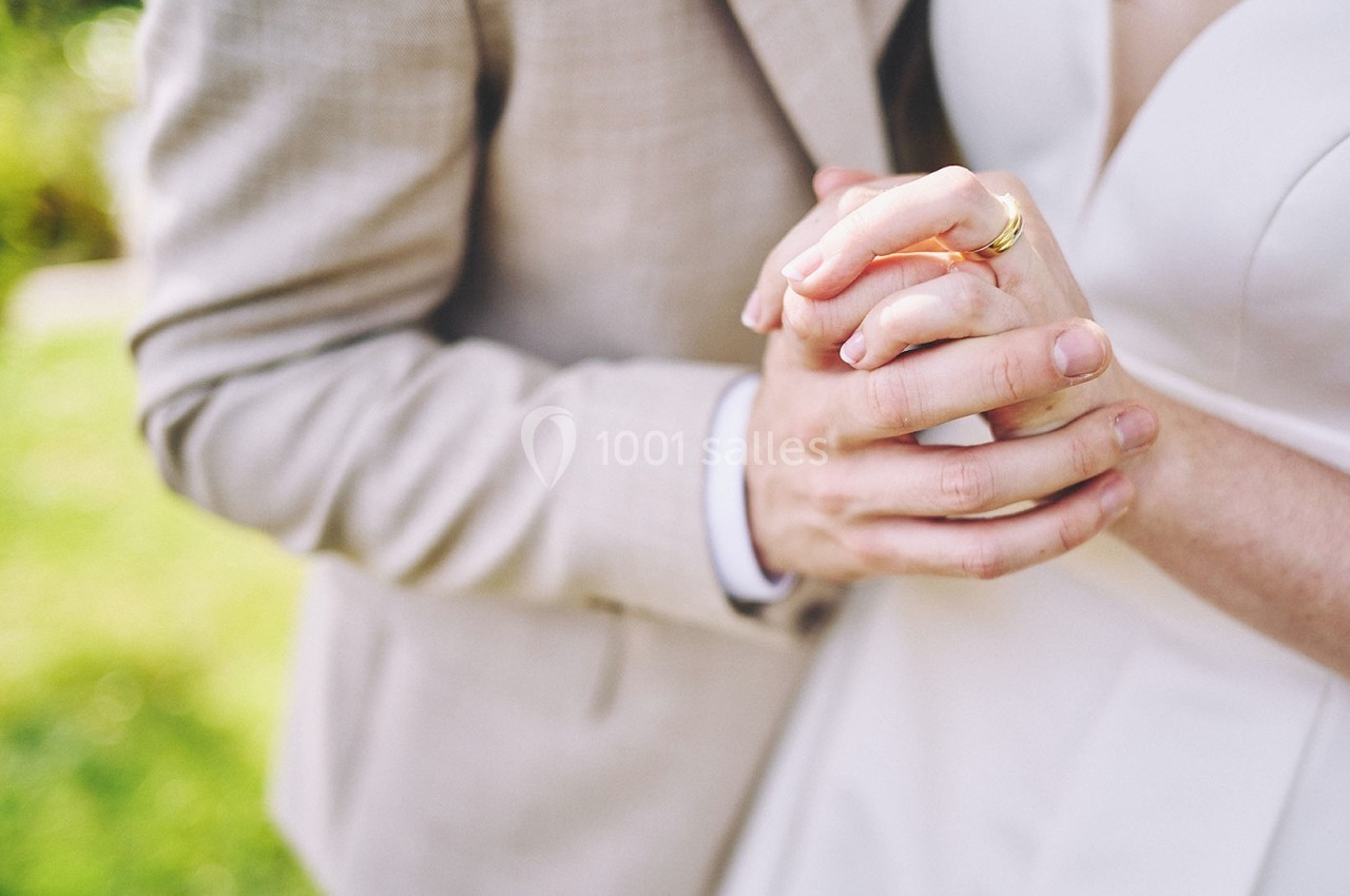 Deux personnes en tenue de mariage se tiennent les mains avec des alliances visibles, dans un cadre extérieur lumineux.