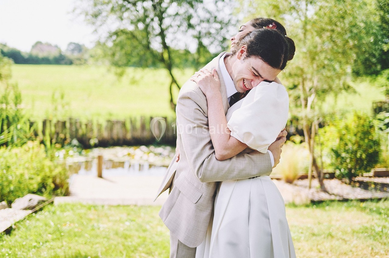 Deux personnes s'enlaçant chaleureusement dans un jardin ensoleillé, avec un étang et des arbres en arrière-plan.