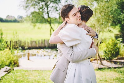 Un couple de mariés s'embrasse tendrement près d'un étang, entouré de verdure, la mariée tenant un bouquet coloré.