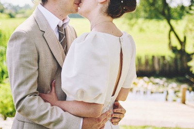 Un couple de mariés s'embrasse tendrement près d'un étang, entouré de verdure, la mariée tenant un bouquet coloré.