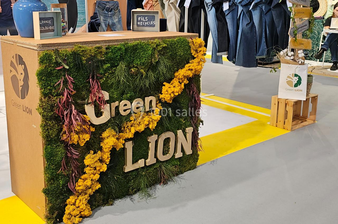 Comptoir décoré de mousse, fleurs jaunes et logo ’Green Lion’, entouré de vêtements en exposition.