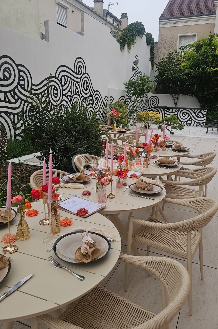 Table extérieure élégamment dressée avec des bougies roses, des fleurs et de la vaisselle, entourée d'un mur décoré de…