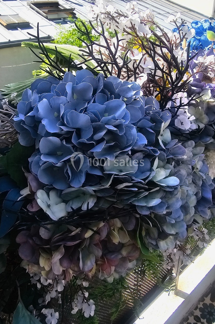 Fleurs artificielles aux teintes bleues et violettes disposées sur un balcon avec vue sur des toits et végétation.