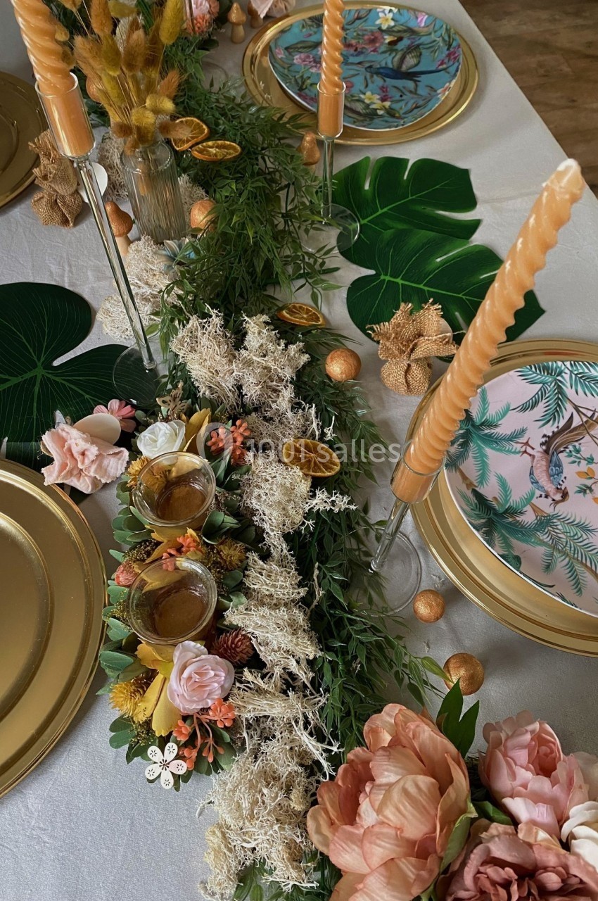 Table décorée avec des fleurs, mousse, feuilles tropicales, bougies torsadées et assiettes aux motifs colorés.