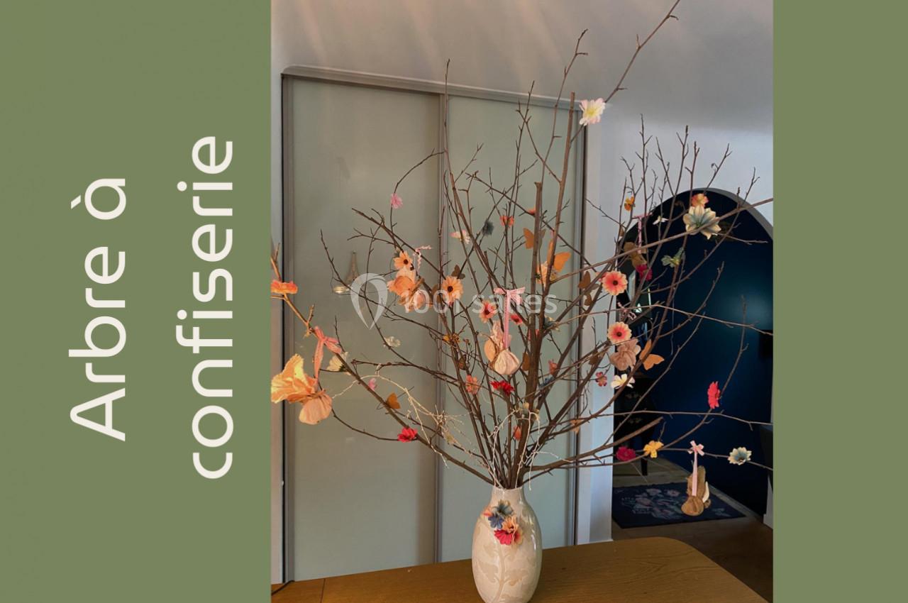 Branches décorées de fleurs en papier coloré dans un vase blanc, posées sur une table en bois devant une armoire.