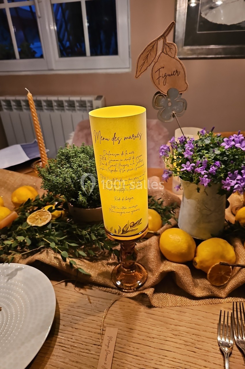Menu manuscrit sur papier jaune posé sur un bougeoir, entouré de citrons, fleurs violettes et décorations de table.