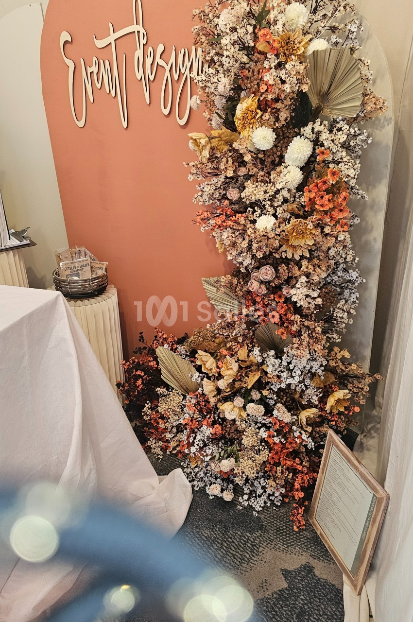Mur décoratif avec fleurs séchées et fraîches dans des tons chauds, accompagné d'une inscription ’Event Design’.