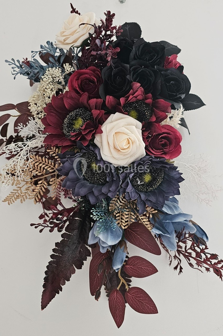 Arrangement floral avec des roses rouges, blanches et noires, des fleurs bleues et des feuillages variés sur fond clair.